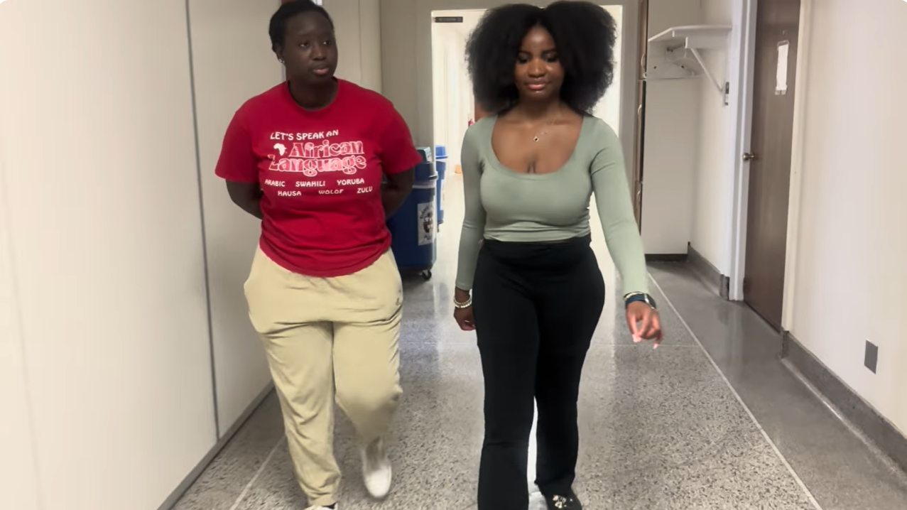 Two students walking side by side along a corridor, one wearing a red T-shirt that reads "Let’s speak an African language".